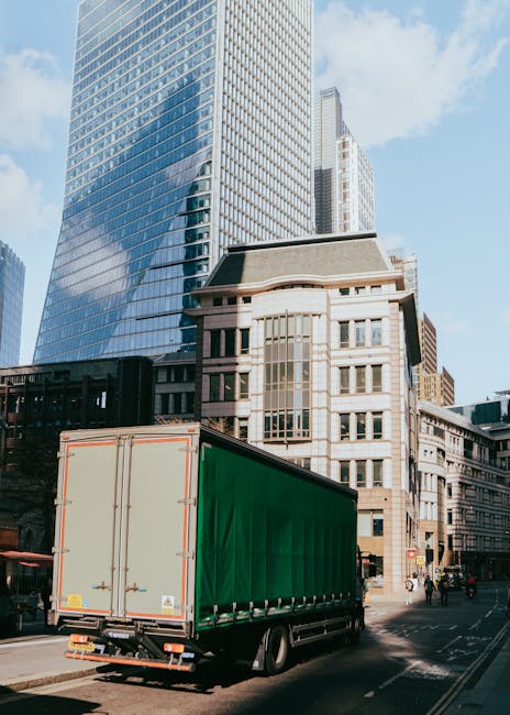 A daytime scene at a coastal location showing a white delivery truck parked on a paved area near the water, with its rear doors closed. To the right, there is a large yellow hydraulic crane positioned
