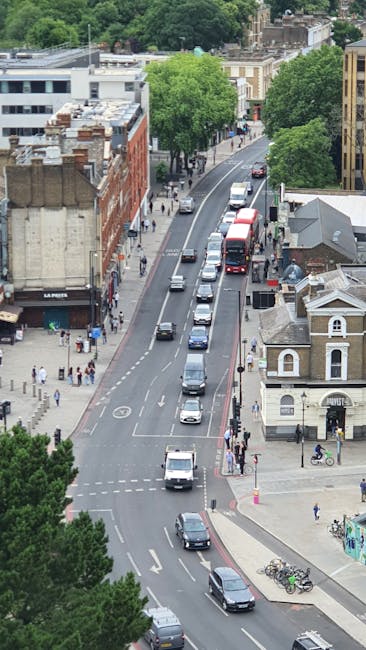 An aerial view of a busy urban street, showing a row of parked cars along the curb, with a variety of moving vehicles including vans and buses traveling in both directions. On the left side, there are