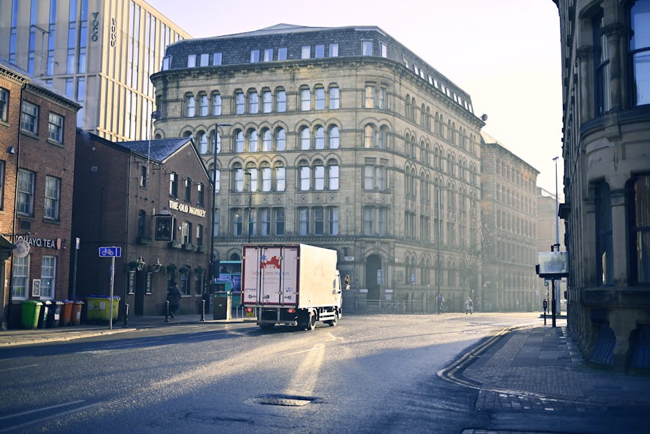 A small box truck with a white cargo area and red logo on its back is parked on a city street near the entrance of a commercial building. The loading process involves the truck positioned close to the